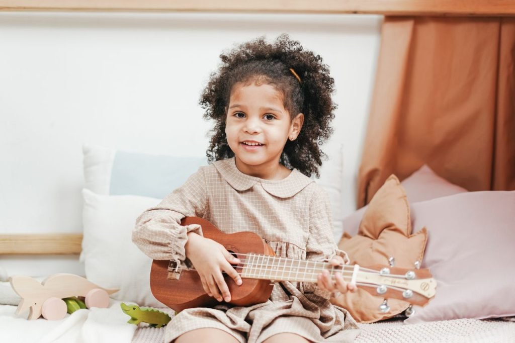 Adorable young girl playing a ukulele while sitting on a bed in a cozy bedroom.